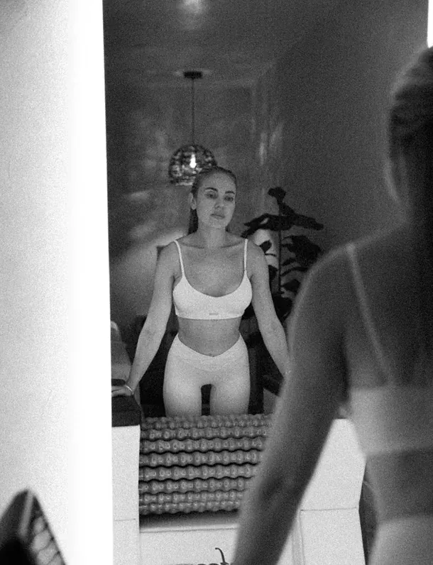 Woman in white activewear by a body roller machine, reflected in the studio mirror