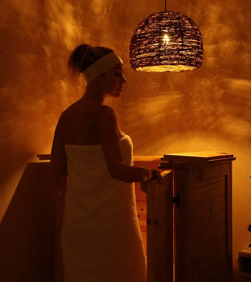 Client wrapped in a towel stepping into the cedar phyto barrel herbal steam sauna under a woven lamp